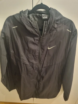 Svart Nike vindjacka med huva - Snygg svart vindjacka från Nike med huva och dragkedja framtill. Jackan har reflekterande detaljer på axlarna och Nike-logga på bröstet.