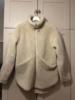 Beige teddyjacka med hög krage - Mysig beige teddyjacka med hög krage och dragkedja framtill. Jackan har en oversized passform, långa ärmar och två stora fickor framtill. Tillverkad i fluffigt teddy-material som håller dig varm under kalla dagar. Perfekt för dig som gillar en avslappnad och trendig stil.