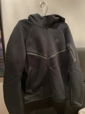Svart Nike hoodie med dragkedja - Säljer en svart hoodie från Nike med dragkedja framtill och huva. Hoodien har en diskret Nike-logga på bröstet och en snygg detalj med en diagonal grå linje över bröstet. Tillverkad i mjukt och bekvämt material. Använd max 3 gånger så riktigt bra skick.