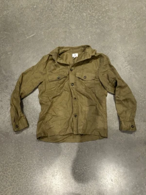 Overshirt - Snygg olivgrön overshirt med två bröstfickor och knappar framtill. Har lång ärm och klassisk krage, perfekt för en avslappnad men stilren look.