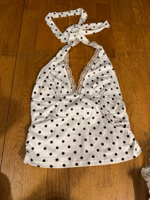Vit prickig halterneck topp XS-S - Säljer en vit halterneck topp med svarta prickar och djup v-ringning dekorerad med spets. Toppen knyts i nacken och har en feminin, retroinspirerad look. Perfekt för dig som vill sticka ut lite extra i sommarvärmen!
