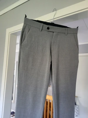Grå kostymbyxor med raka ben - Snygga grå kostymbyxor med raka ben och pressveck. Byxorna har hällor för bälte, sidfickor och stängs med knapp och dragkedja. Perfekta för en clean och stilren look.