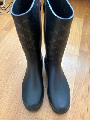 Svarta gummistövlar från Anna Field - Snygga svarta gummistövlar från Anna Field med klassiskt monogrammönster upptill. Stövlarna är höga och har en stilren, matt finish. Perfekta för regniga dagar när du vill hålla stilen. Tillverkade i slitstarkt gummi och har en diskret knappdetalj vid skaftet. Helt nya och oanvända med prislapp kvar. Köpta för 399, säljer för 350. Storlek 40. 