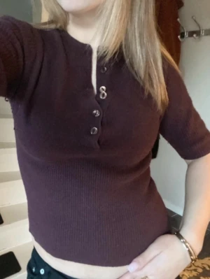 Mörklila/vinröd  ribbad topp med knappar - Trendig mörklila/vinröd ribbad topp med korta ärmar och knappdetaljer framtill. Toppen har en figurnära passform och är perfekt för att skapa en snygg och enkel look.