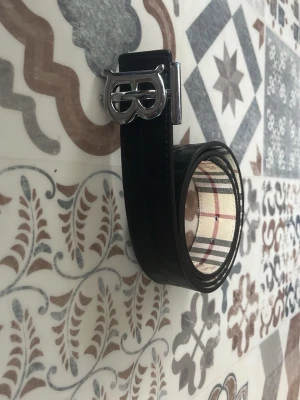 Svart bälte med silverfärgat spänne - Snyggt svart bälte från Burberry med klassiskt rutigt mönster på insidan och ett stilrent silverfärgat spänne med logga. Bältet har en elegant och tidlös design som passar till många olika outfits.