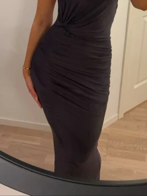 Balklänning - Fest klänning  - Snygg mörk långklänning med tight passform och draperade detaljer. Klänningen har en elegant öppen rygg och är ärmlös, vilket ger en stilren och modern look. Perfekt för dig som vill sticka ut med en sofistikerad stil.