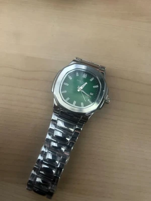 Silverfärgad klocka med grön urtavla - Stilren klocka från Nautica med grön urtavla och datumvisning. Klockan har ett robust armband i silverfärgad metall och klassisk rund boett. Perfekt accessoar för dig som vill sticka ut med en snygg detalj på handleden.