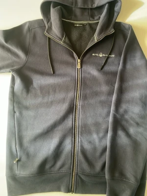 Svart hoodie från Sail Racing - Svart hoodie med dragkedja från Sail Racing. Klassisk modell med huva och dragsnören, två fickor framtill och logga på bröstet samt på huvan. Tillverkad i mjukt och bekvämt material, perfekt för en avslappnad stil. Pris kan diskuteras!!!!