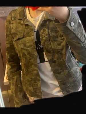 Camo jeansjacka i grön nyans - Snygg jeansjacka med camouflage-mönster i gröna och beige toner. Klassisk modell med knappar framtill och bröstfickor. Perfekt för dig som vill sticka ut med en cool streetstyle-look.