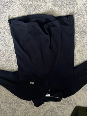 Mörkblå henley från Lacoste - Snygg mörkblå henley från Lacoste med klassisk krage och knappar framtill. Ikoniska krokodilloggan på bröstet. Perfekt för en clean och stilren look. Tillverkad i mjukt bomullsmaterial som känns skönt mot huden.