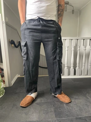 Svarta cargobyxor YA med fickor - Snygga svarta cargobyxor från YA med flera stora fickor på sidorna och dragsko i midjan. Byxorna har en avslappnad passform och är perfekta för en streetwear-look. Materialet ser ut att vara mjukt och bekvämt, perfekt för chill dagar.