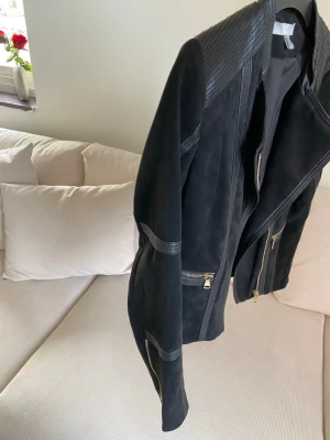 Svart PU-mockajacka med dragkedjor - Snygg svart PU-mockajacka från HM med coola detaljer som dragkedjor i silver och ribbade partier på axlar och ärmar. Jackan har en asymmetrisk dragkedja framtill och flera fickor med dragkedja. Perfekt för en edgy stil.