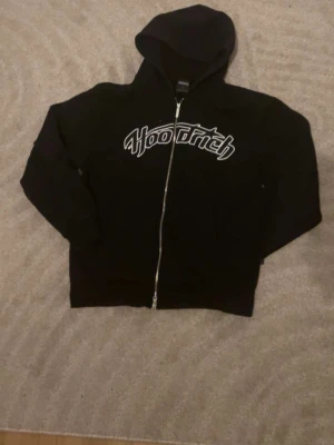 Svart Hoodrich hoodie - Säljer en svart hoodie från Hoodrich med dragkedja framtill och stor vit logga på bröstet. Hoodien har huva och långa ärmar