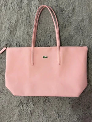 Ljusrosa shoppingväska från Lacoste - Snygg ljusrosa shoppingväska från Lacoste med klassisk krokodillogga framtill. Väskan har två långa handtag och dragkedja upptill. Tillverkad i slätt syntetmaterial som ger en stilren look. Perfekt för att bära allt du behöver under dagen. Helt ny aldrig använt men har bara en liten fläck som syns inte mycket ( sista bilden).orginalspris 1245 kr