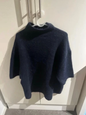 Mörkblå stickad poncho - Mysig mörkblå stickad poncho med hög krage och vid passform. Perfekt att slänga på sig över en tröja för extra värme. Enkel och stilren design utan synliga detaljer eller mönster.