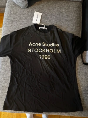 Acne studios t shirt - Cool svart t-shirt från Acne Studios med vit text 'Acne Studios STOCKHOLM 1996' på bröstet. Klassisk passform och rund halsringning. Perfekt för dig som gillar stilrena och trendiga plagg. Modellen är oversize