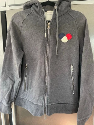 Grå Moncler hoodie med dragkedja - Mörkgrå/blå hoodie från Moncler med dragkedja framtill, två fickor med dragkedja och huva med dragsko. Bröstet har Monclers ikoniska logga i rött, vitt och blått. Insidan är fodrad med mjuk fleece för extra komfort. Perfekt för en avslappnad och snygg stil. Storlek är M, något liten i storlek 