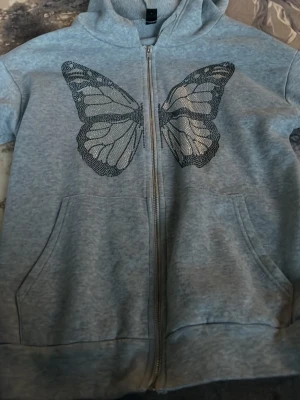 Grå hoodie med fjäril från SHEIN - Grå zip-hoodie från SHEIN med stor svart fjäril framtill. Hoodien har huva, dragkedja och två fickor. Mjuk insida och avslappnad passform, perfekt för en chill look.