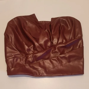 Brun tubtopp med dragkedja bak - Snygg brun tubtopp i skinnimitation med cool drapering framtill och dragkedja bak. Toppen är axelbandslös och har en croppad passform som passar perfekt till höga jeans eller kjol. Storlekslappen är borttagen men skulle säga strl S. Katt finns i hemmet.