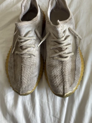Beige Yeezy Boost 350 sneakers - Säljer ett par beige Yeezy Boost 350 sneakers med stickad ovandel och klassisk ribbad sula i gulbrunt gummi. Skorna har rund tå, snörning och en diskret rand på sidan. Perfekta för dig som gillar streetwear och vill ha något bekvämt och stilrent.