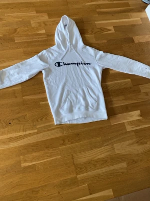 Vit Champion hoodie med logga - Säljer en vit hoodie från Champion med stor svart logga på bröstet och liten logga på ärmen. Hoodien har huva med dragsko och en klassisk magficka. Tillverkad i mjukt bomullsmaterial som är skönt att bära. Perfekt för en avslappnad och sportig stil.