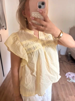 Gul blus med volangärmar från studio - Söt ljusgul blus från studio med volangiga korta ärmar och broderade detaljer. Blusen har en lätt v-ringning med liten volangkant och är tillverkad i ett tunt, luftigt tyg med diskret prickstruktur. Perfekt för en somrig och fräsch look.