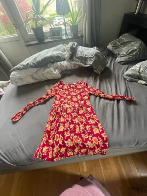 Röd blommig långärmad klänning - Fin röd klänning med blommigt mönster i gult och beige. Klänningen har lång ärm, rund halsringning och ett lätt utställt kjolparti. Perfekt för dig som gillar färg och mönster!