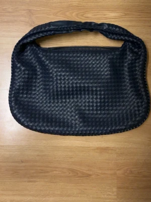 Mörkblå flätad handväska i skinn från Design by si - Snygg svart handväska med flätad design och mjuk, rundad form. Väskan har ett kort handtag och är tillverkad i skinn. Köpt ifrån Design by si för 605kr. Använd men i bra skick, saknar dock läder-handtaget för dragkedjan men annars hel går såklart att stänga ändå. Väldigt rymlig fin väska säg till för fler bilder! 