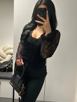 Svart spetsblus med lång ärm - Snygg svart blus i spets med långärmade transparenta ärmar och knappar framtill. Figurnära modell med v-ringning och fina spetsdetaljer som ger en trendig look. Perfekt att styla med svarta byxor eller kjol.