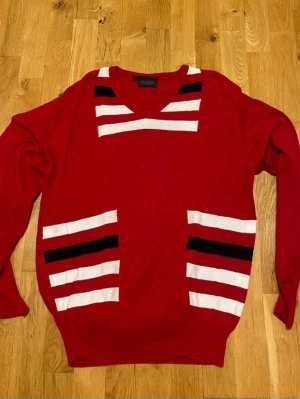 Röd stickad tröja från Dahlin stl L - Röd stickad tröja från Dahlin med vita och svarta ränder på bröstet och framtill. Tröjan har V-ringning och långa ärmar. Perfekt för dig som gillar färgstarka och unika plagg med sportig känsla.