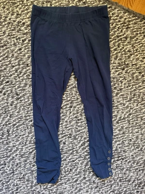 Mörkblå leggings med knappar - Mörkblå leggings i mjukt stretchigt tyg med bred resår i midjan. Nedtill på benen finns dekorativa knappar och lätt rynkade detaljer. Perfekta för en avslappnad och trendig look. Perfekt under kjol/klänning.