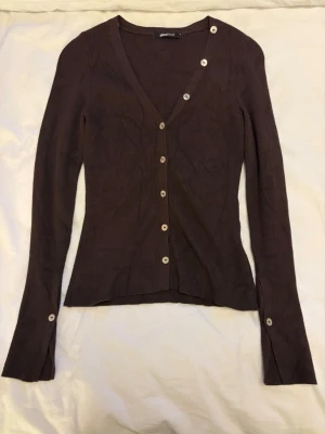Brun ribbad kofta från Gina Tricot - Snygg brun ribbad kofta från Gina Tricot med v-ringning och vita knappar hela vägen fram. Långärmad modell med extra knappar vid ärmsluten. Perfekt att styla lager på lager.
