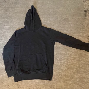 Svart hoodie med magficka - En klassisk svart hoodie med stor huva och rymlig magficka framtill. Hoodien har ribbade muddar vid ärmslut och nederkant. Perfekt för en avslappnad och stilren look. Materialet känns mjukt och bekvämt, troligen bomullsblandning. Storlek 158/164.
