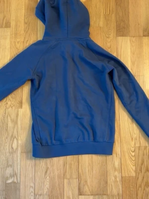 Blå hoodie från Peak Performance - Mörkblå hoodie med vit Peak Performance-logga på bröstet. Klassisk modell med huva, dragsnören och stor magficka framtill. Tillverkad i mjukt bomullsmaterial och har ribbade muddar vid ärmslut och nederkant. Perfekt för en avslappnad och sportig stil. 