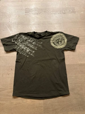 Grön t-shirt med tryck - Grön t-shirt med stort Medusa-tryck på bröstet och flera loggor i vitt över vänster sida. Klassisk rund hals och korta ärmar. Tillverkad i mjuk bomull för skön känsla och snygg passform.