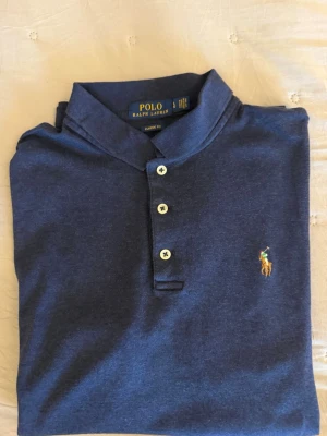 Marinblå soft touch pikétröja från Ralph Lauren - Klassisk marinblå pikétröja från Ralph Lauren med krage och tre knappar framtill. Tröjan har det ikoniska broderade polospelar-loggan på bröstet och är i en skön bomullsblandning. Perfekt för en stilren och avslappnad look.