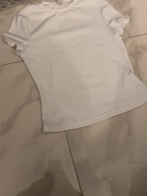 Vit basic t-shirt träningsmatriall - En klassisk vit t-shirt med rund hals och korta ärmar. Enkel och stilren design, perfekt att matcha med jeans eller shorts. Tillverkad i mjuk bomull för en bekväm känsla hela dagen.