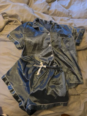 Silvrig satinpyjamas med kort ärm - Snygg tvådelad pyjamas i silvrig satin med kortärmad skjorta och matchande shorts. Skjortan har vita knappar, bröstficka och vit passpoal. Shortsen har resår i midjan och vit rosett. Perfekt för mysiga hemmakvällar.