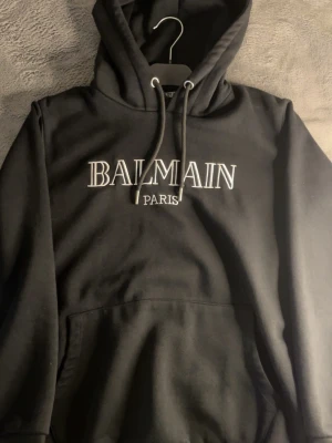 Svart Balmain hoodie med tryck - Snygg svart hoodie från Balmain med vit broderad logga 'BALMAIN PARIS' på bröstet. Klassisk modell med huva och dragsnören samt en stor magficka. Mjuk bomullskänsla och stilren design som passar perfekt till streetwear.