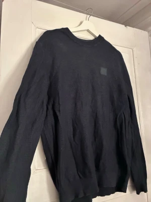 Svart tröja från BOSS med struktur - Säljer en svart långärmad tröja från BOSS med diskret logotyp på bröstet. Tröjan har en strukturerad, stickad känsla och rund halsringning. Perfekt för dig som gillar stilrena och klassiska plagg med en modern touch. Passar snarare M/L.