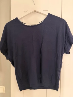 Mörkblå topp med spetsdetalj - Mörkblå topp med lös passform och korta ärmar. Baksidan har en unik spetsdetalj i virkad stil som ger toppen en cool twist. Perfekt till jeans eller kjol för en avslappnad look.