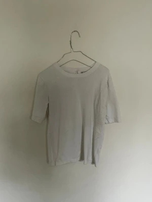 Vit enkel kortärmad topp - Vit basic topp med rund halsringning och korta ärmar. Tillverkad i mjuk bomull, perfekt för en clean och stilren look. Liten knappdetalj i nacken för enkel påklädning. Passar till allt och är ett måste i garderoben.