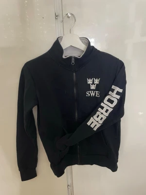 Rid Svart zip-hoodie  - Svart ridtröja med dragkedja och hög krage. På bröstet finns ett vitt SWE-tryck med kronor och på ena ärmen står det HORSE i stora bokstäver. Muddar vid ärmslut och nederkant. Perfekt för dig som gillar hästar och sportig stil.