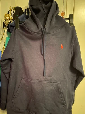 Mörkblå hoodie från Polo Ralph Lauren - Mörkblå hoodie från Polo Ralph Lauren med klassisk huva och dragsko. Har en röd broderad logga på bröstet och en stor känguruficka framtill. Perfekt för en avslappnad och stilren look. Tillverkad i mjukt bomullsmaterial.