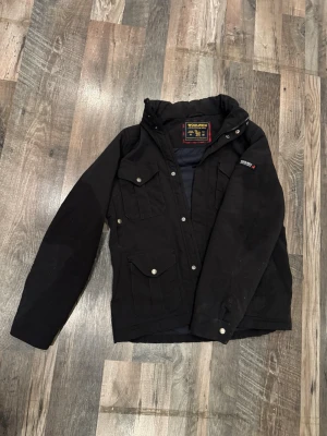Svart jacka från Woolrich - Snygg svart jacka från Woolrich. Extra eftertraktad då jackan inte tillverkas längre. Skick 4/5 då den har andvända lite men det finns inga defekter eller liknande. Jackan har tryckknappar, dragkedja och logga på ärmen. Skriv privat för flera bilder eller om du har frågor.