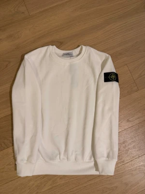 Vit sweatshirt från Stone Island - Vit sweatshirt från Stone Island med klassisk rund hals och lång ärm. Tröjan har den ikoniska svarta logotyp-patchen på vänster ärm. Perfekt för en clean och stilren look.