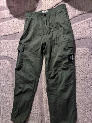 Olivgröna cargobyxor Calvin Klein - Snygga olivgröna cargobyxor från Calvin Klein Jeans med elastisk midja och flera praktiska fickor på sidorna. Byxorna har en klassisk cargomodell och en tydlig CK-logga på ena benet. Perfekta för en avslappnad och trendig stil.
