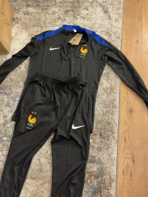 Frankrike Nike träningsset svart/blå - Snyggt träningsset från Nike med Frankrikes landslagsemblem. Setet består av en svart långärmad tröja med blå detaljer på axlarna och dragkedja vid halsen samt matchande svarta byxor. Båda plaggen har Nike-logga och FFF-märke med tupp och stjärnor. Perfekt för fotbollsträning.