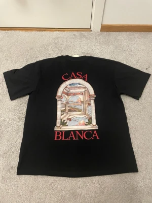 Svart Casa Blanca t-shirt med tryck - Svart t-shirt från Casablanca med färgglatt tryck av en portal och landskap på ryggen samt mindre tryck på bröstet. Röd text 'Casa Blanca' både fram och bak. Klassisk passform och rund hals. Perfekt för dig som gillar unika prints.