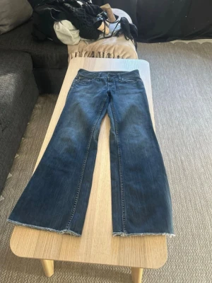 Blå bootcut jeans med rå kant - Säljer ett par blå bootcut jeans med rå, fransad kant nertill. Jeansen har klassisk femficksdesign och låg midja. Perfekta för dig som gillar en avslappnad och trendig look. Materialet är denim i bomull. 